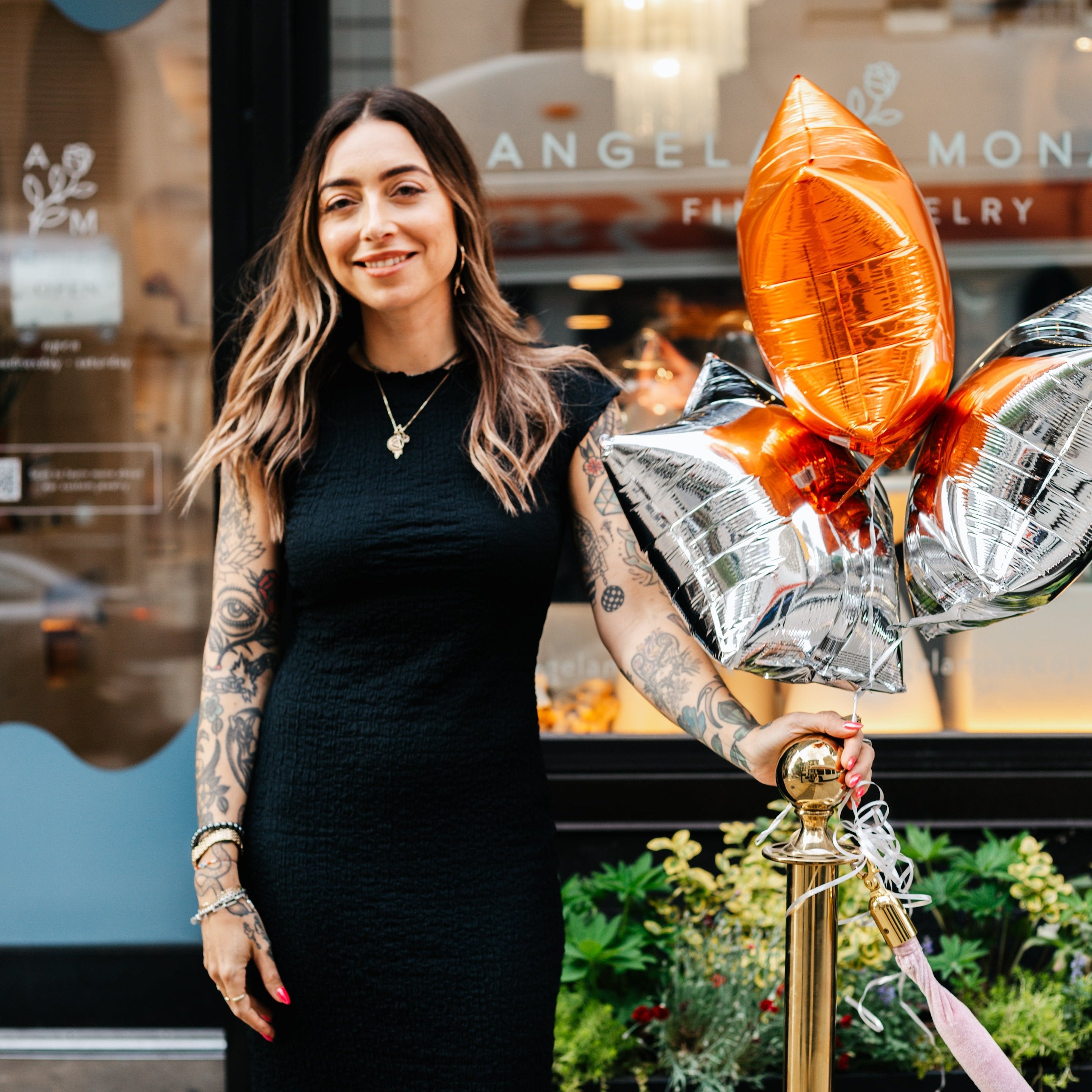 Photo of Angela Monaco our founder wearing a sleeveless black dress, standing next to a bouquet of multicolored balloons in front of our Rittenhouse flagship store 