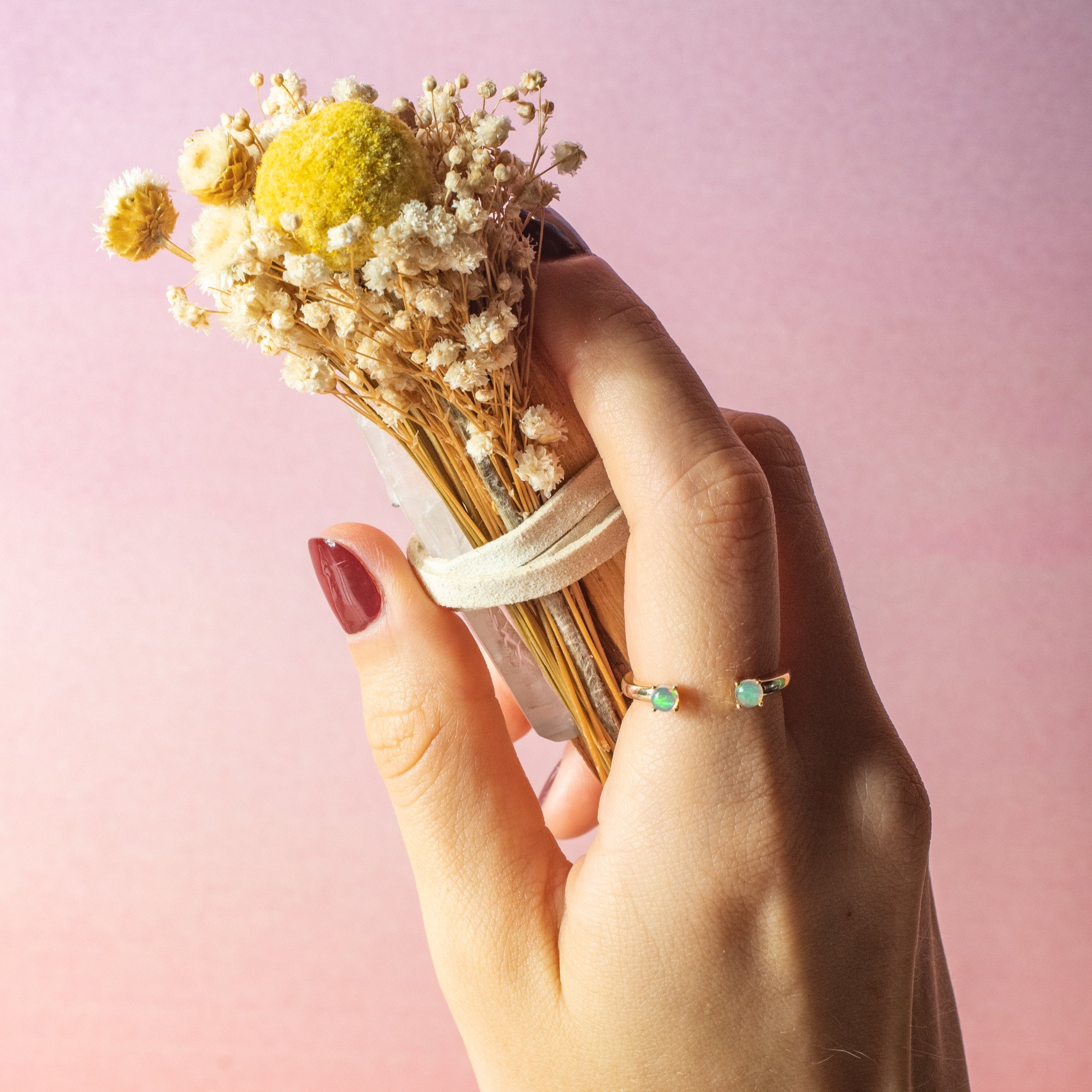A close-up of a hand holding the PALO SANTO & QUARTZ BUNDLE  by BLACK AND JANE . The bundle features small white blossoms and a prominent yellow billy button flower. The hand, with dark red nails, wears a gold ring adorned with two small green gemstones. The background is a soft pink gradient, creating a warm and elegant composition.