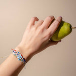 Hand holding green pear on beige background wearing all colors of black and jane evil eye bracelets including black, red, blue pink, teal and white