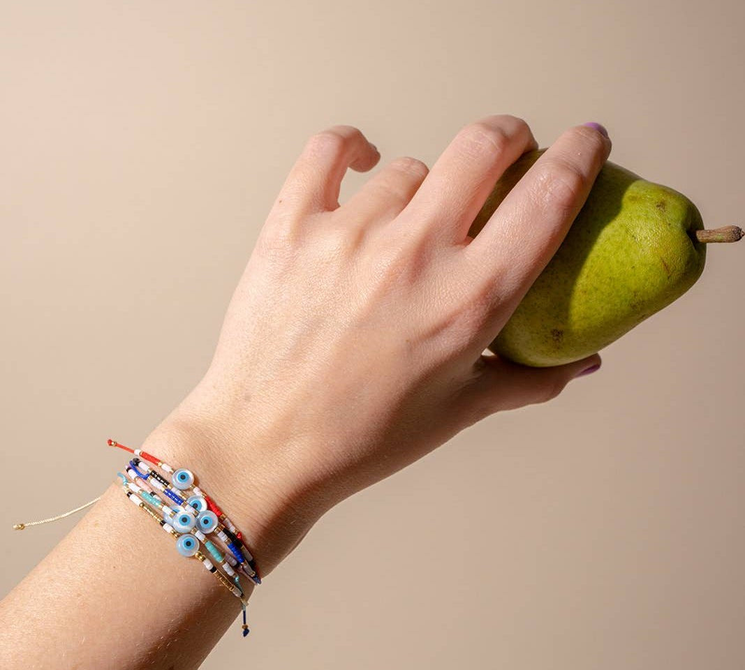 Hand holding green pear on beige background wearing all colors of black and jane evil eye bracelets including black, red, blue pink, teal and white